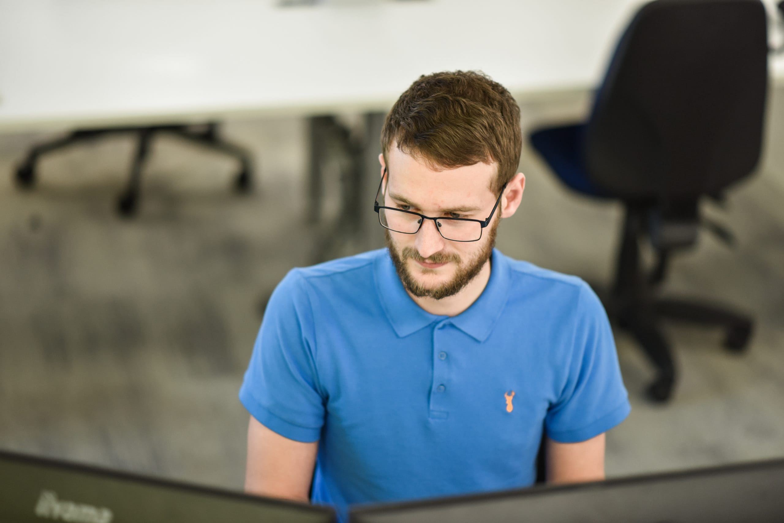 Photograph of a Cantium employee in a blue shirt and glasses, sat behind computer screens while working on analytics.