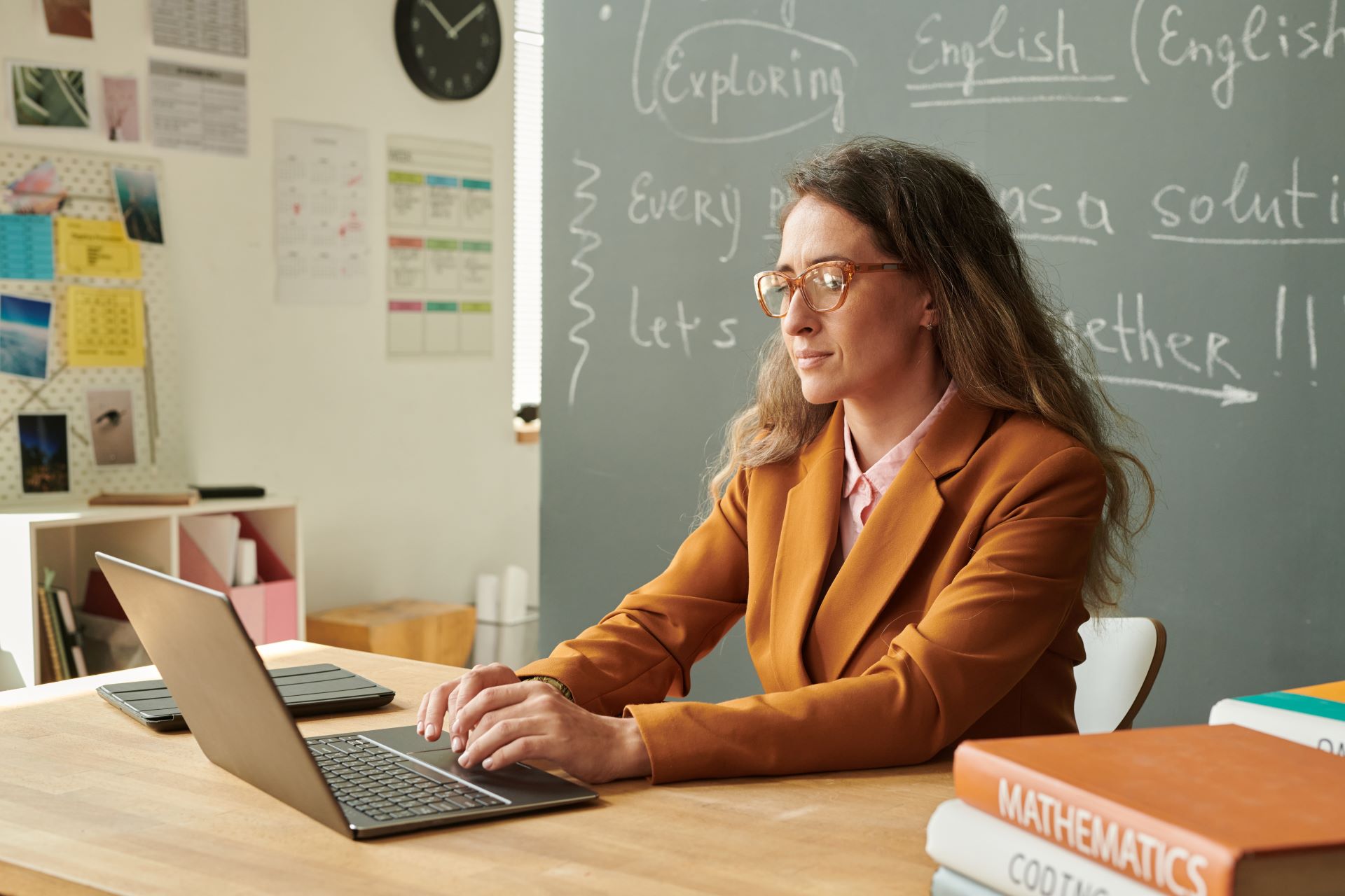 Teacher sitting at desk typing on laptop with chalkboard in the background.