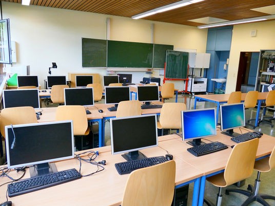 Photograph of a classroom. Each desk has a computer, monitor and keyboard on it. There is nobody in the room.
