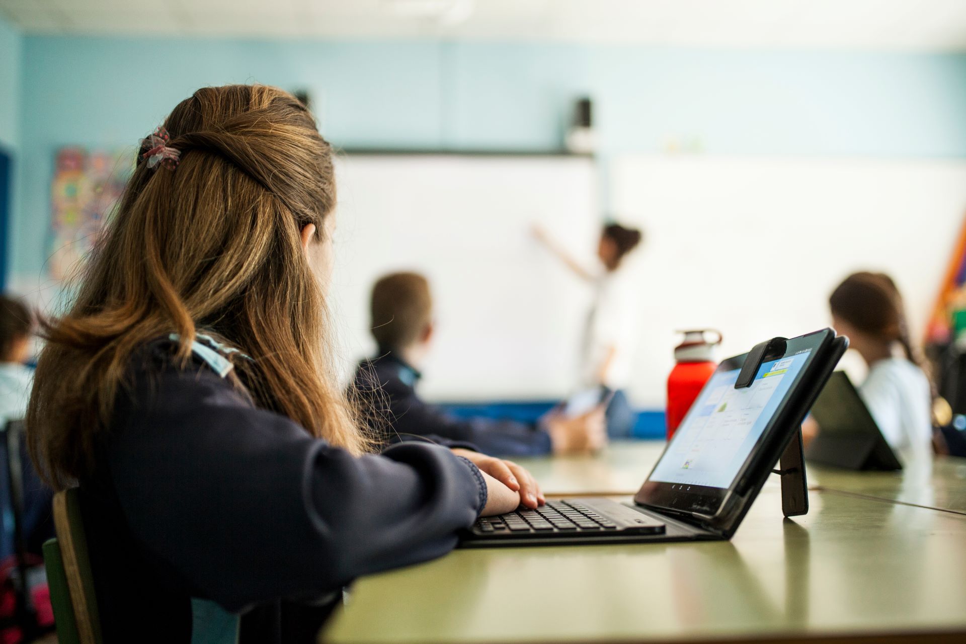 Girl in school uniform sitting at a table in a classroom with an open tablet in front of her looking at teacher who is writing on a white board.