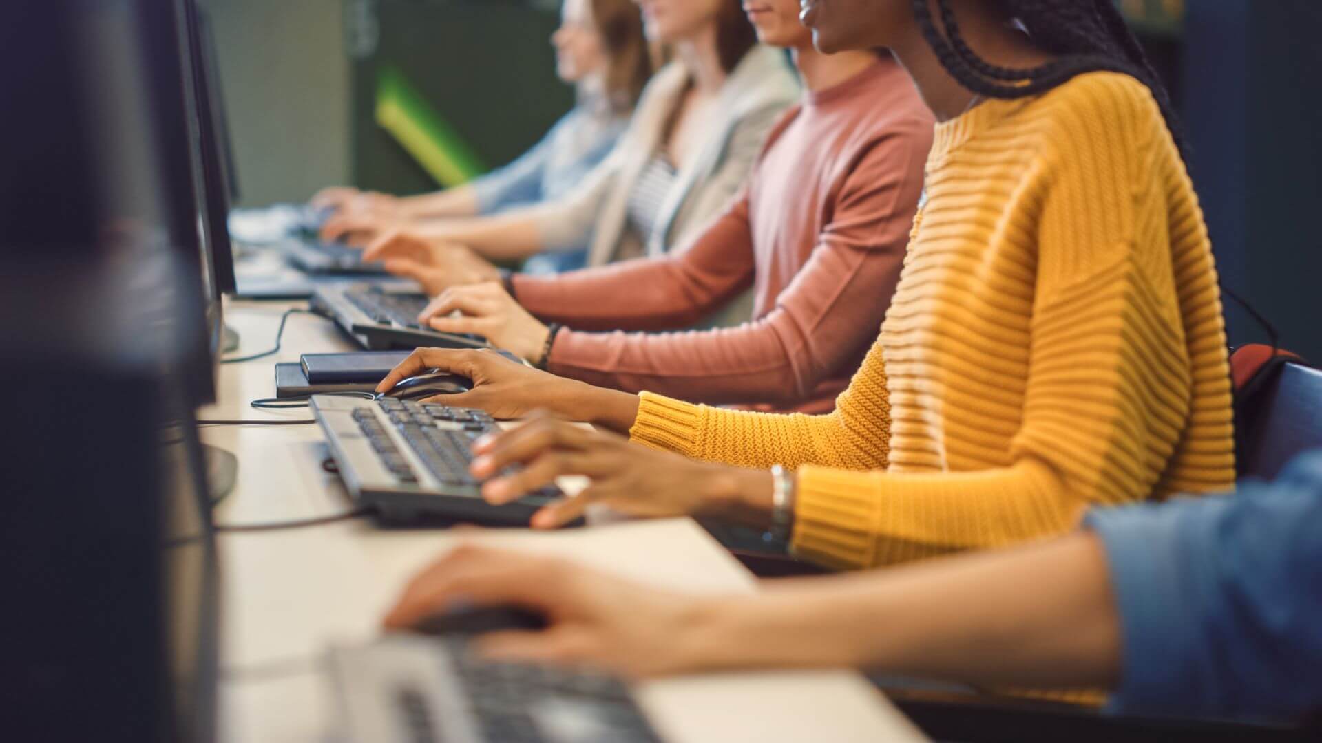 Close up of give people working at a long workstation with computer screens in front of them and hands on keyboards