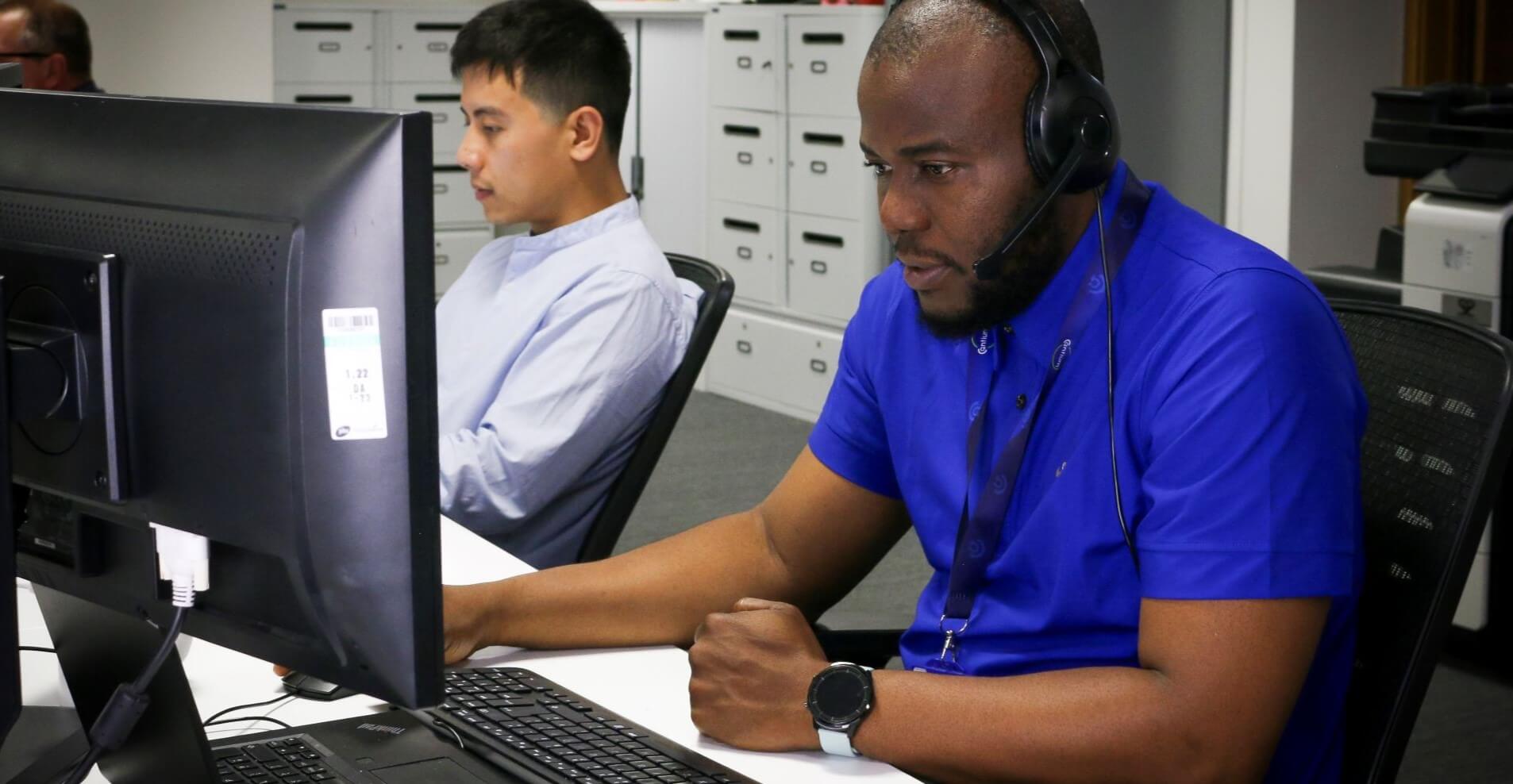 Two people sitting behind computer screens. Person in foreground wearing a headset.